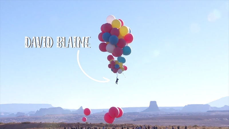 Man is suspended in the air by a clump of large balloons over a crowd of people. There are multiple desert towers and rock features in the distance behind them. Text on screen reads, "David Blaine," with an arrow pointing to the man.