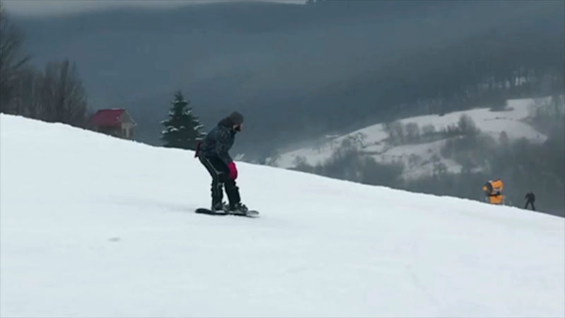 Man wearing a gray winter hat, a dark gray hoodie, black snow pants, and red gloves snowboards down a snowy ski slope on a foggy day. A yellow snow making machine and a little building with a red roof are nearby.