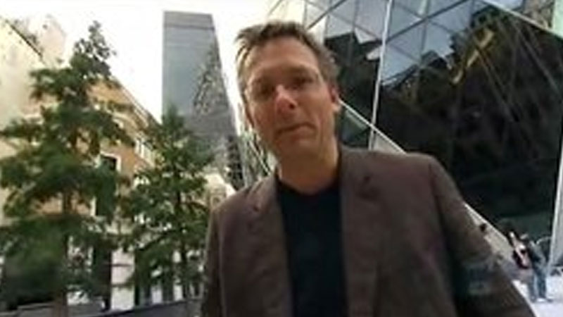 Low angle shot of a man in a suit and glasses standing in front of tall buildings.